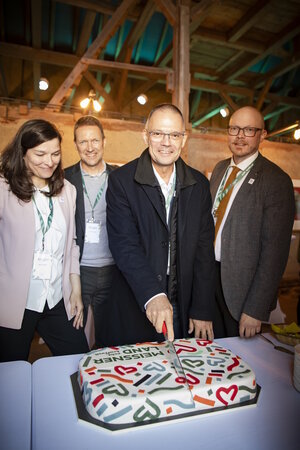 Bild vergr&ouml;&szlig;ern: Vier Personen stehen in einer Scheune vor einem Kuchen mit dem Logo des MEISSNER LAND. Landrat Ralf H&auml;nsel schneidet den Kuchen an.