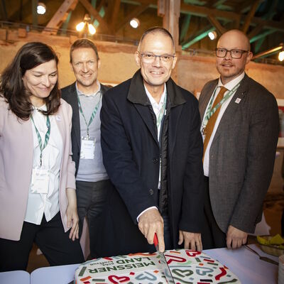 Vier Personen stehen in einer Scheune vor einem Kuchen mit dem Logo des MEISSNER LAND. Landrat Ralf H&auml;nsel schneidet den Kuchen an.
