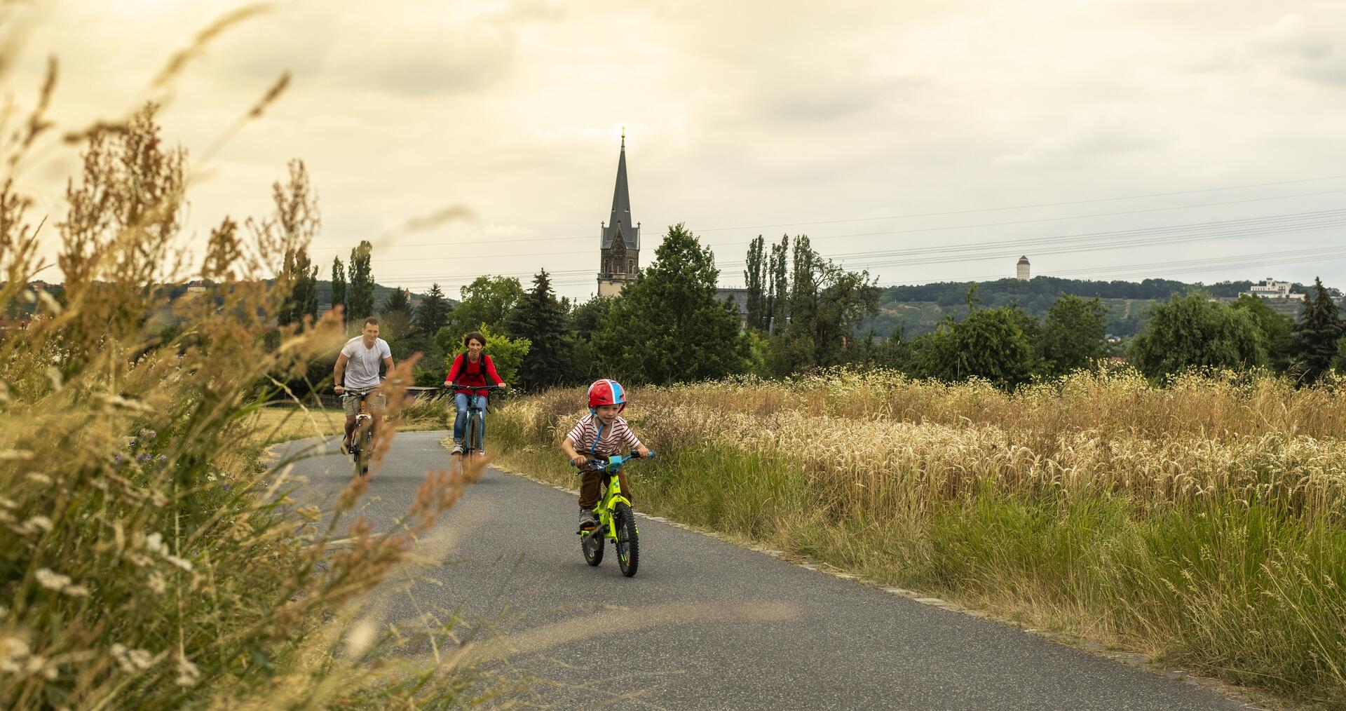 Bild vergrößern: Eine Familie fährt auf einem Radweg Fahrrad. Im Hintergrund eine Kirchturmspitze.